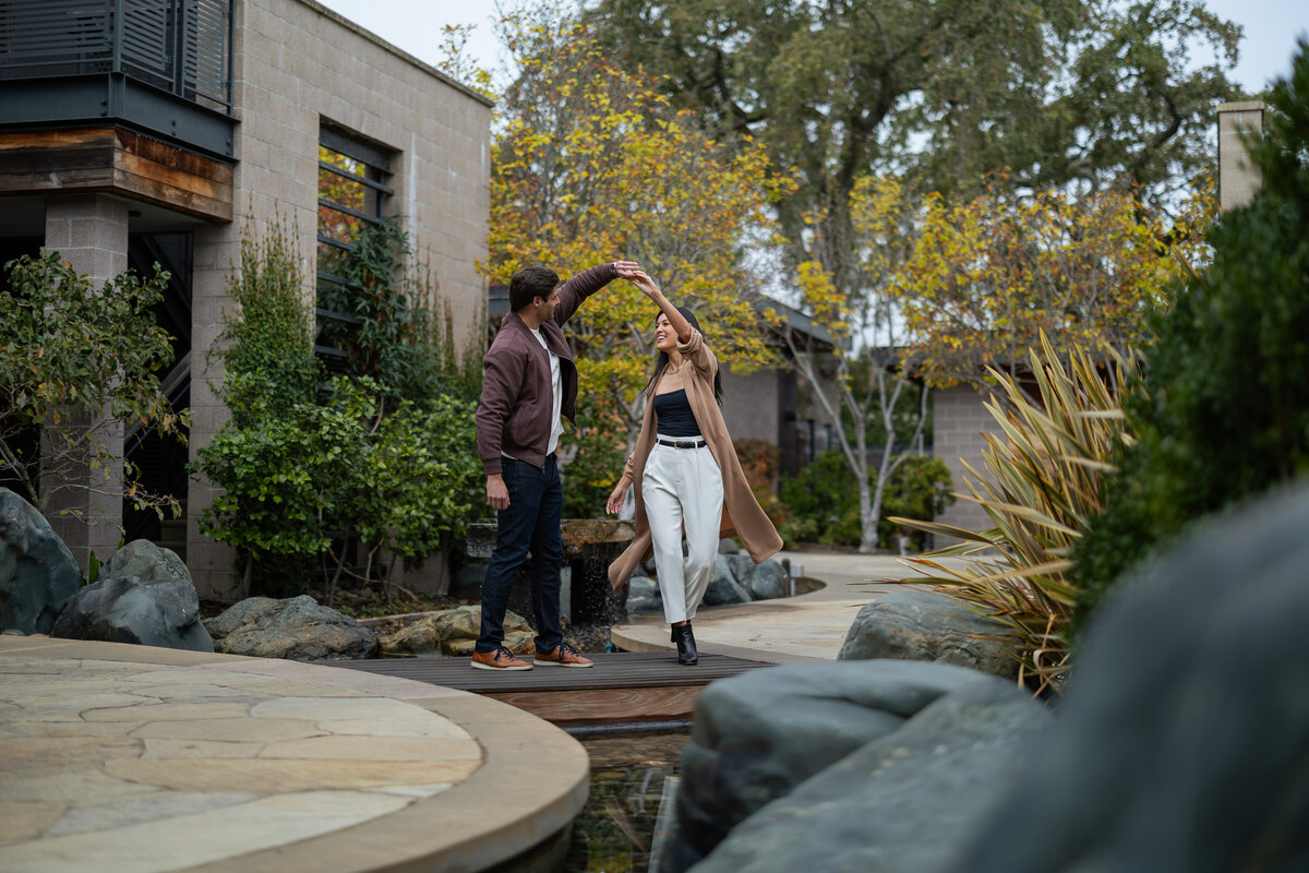 A couple dancing on a bridge in a landscaped outdoor area with a building in the background.
