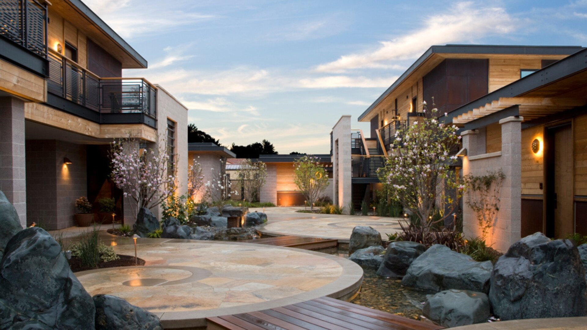A serene courtyard with modern buildings, landscaping, and a water feature at dusk.
